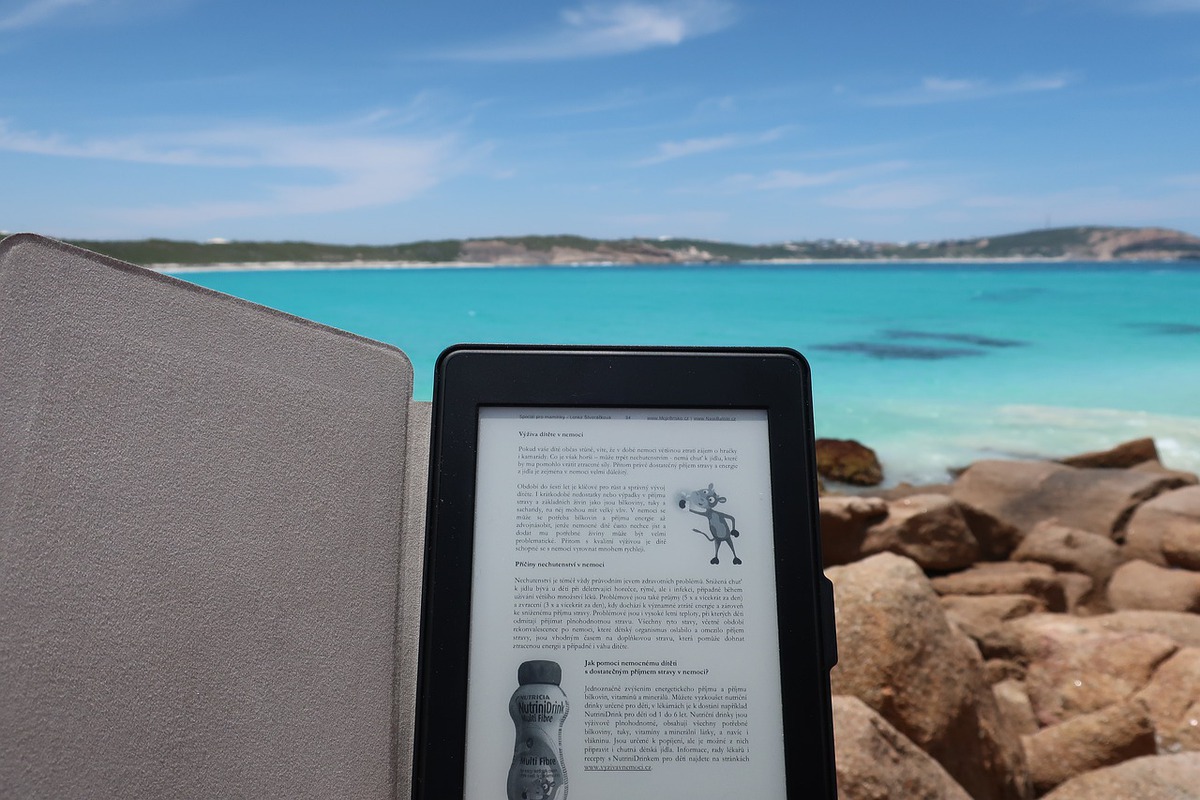  The image shows an Android smartphone with a book open on it. The smartphone is sitting on a beach with the ocean in the background.